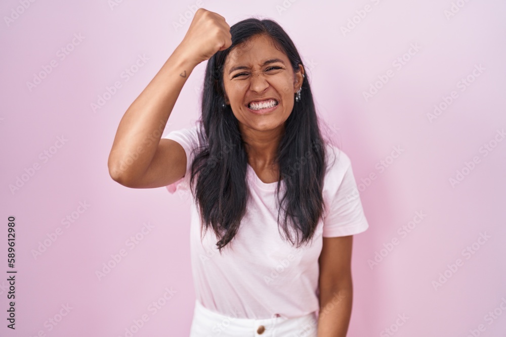 Young hispanic woman standing over pink background angry and mad raising fist frustrated and furious while shouting with anger. rage and aggressive concept. Young hispanic woman standing over pink background angry and mad raising fist frustrated and furious while shouting with anger. rage and aggressive concept.