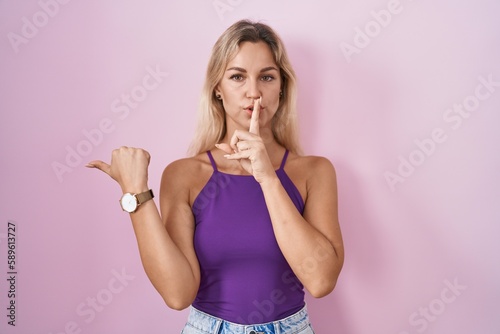 Young blonde woman standing over pink background asking to be quiet with finger on lips pointing with hand to the side. silence and secret concept.