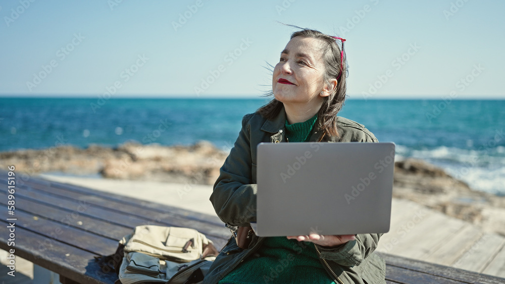 Mature hispanic woman with grey hair tourist using laptop sitting on bench at seaside