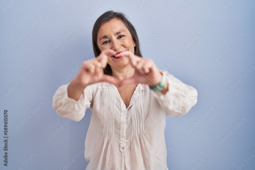 Middle age hispanic woman standing over blue background smiling in love doing heart symbol shape with hands. romantic concept. Middle age hispanic woman standing over blue background smiling in love doing heart symbol shape with hands. romantic concept.
