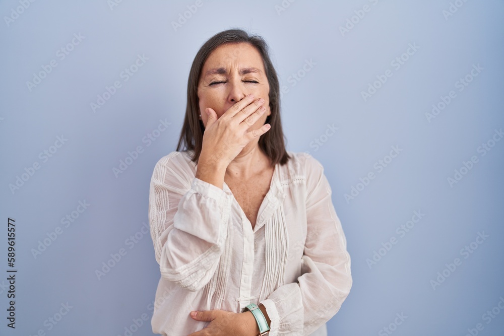 Middle age hispanic woman standing over blue background bored yawning tired covering mouth with hand. restless and sleepiness. Middle age hispanic woman standing over blue background bored yawning tired covering mouth with hand. restless and sleepiness.