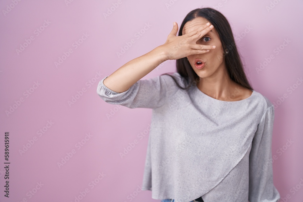 Young brunette woman standing over pink background peeking in shock covering face and eyes with hand, looking through fingers with embarrassed expression. Young brunette woman standing over pink background peeking in shock covering face and eyes with hand, looking through fingers with embarrassed expression.