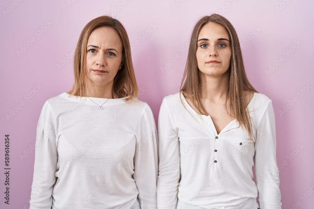 Middle age mother and young daughter standing over pink background relaxed with serious expression on face. simple and natural looking at the camera. Middle age mother and young daughter standing over pink background relaxed with serious expression on face. simple and natural looking at the camera.
