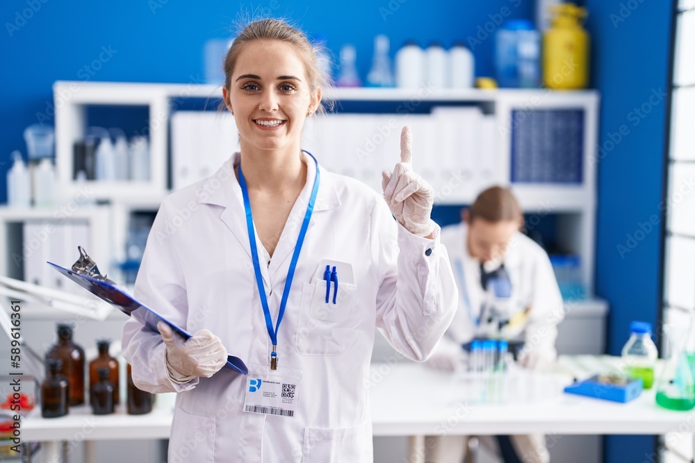 Blonde woman working at scientist laboratory surprised with an idea or question pointing finger with happy face, number one