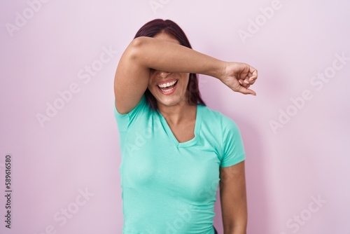 Young hispanic woman standing over pink background covering eyes with arm smiling cheerful and funny. blind concept.