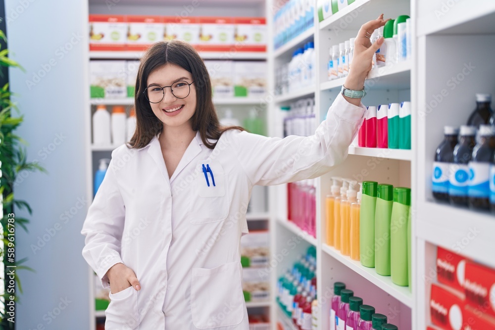 Young caucasian woman pharmacist smiling confident holding medicine on shelving at pharmacy