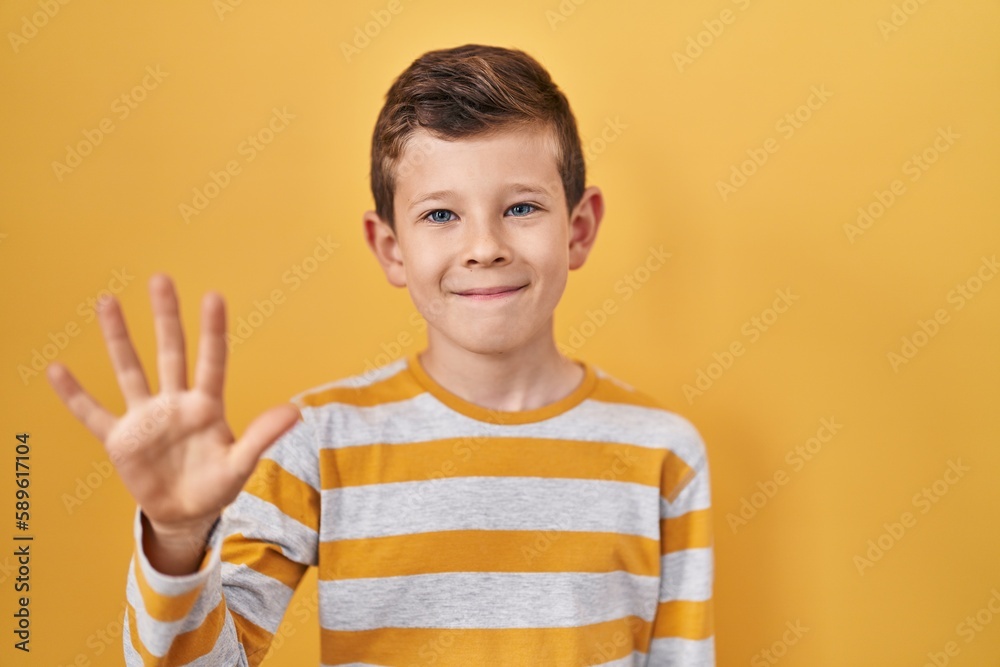 Young caucasian kid standing over yellow background showing and pointing up with fingers number five while smiling confident and happy.