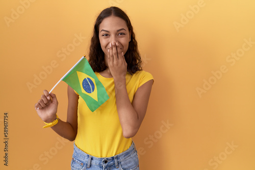 Young hispanic woman holding brazil flag laughing and embarrassed giggle covering mouth with hands  gossip and scandal concept
