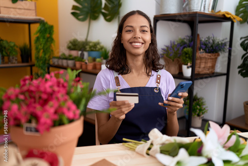 Young african american woman florist using smartphone holding credit card at flower shop