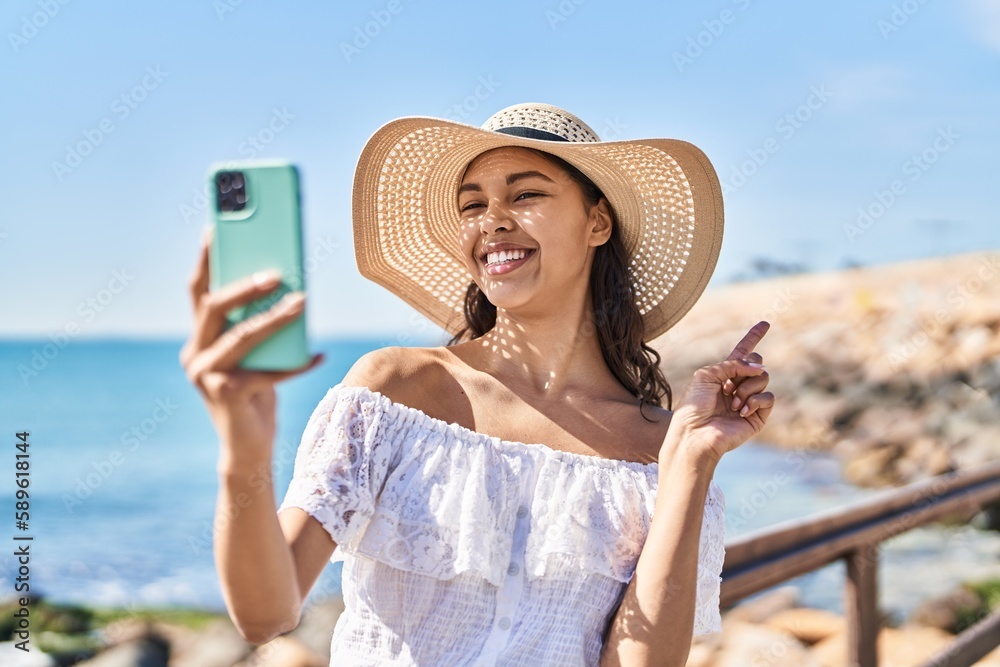 Fototapeta premium Young brazilian woman taking a selfie photo with smartphone outdoors smiling happy pointing with hand and finger to the side