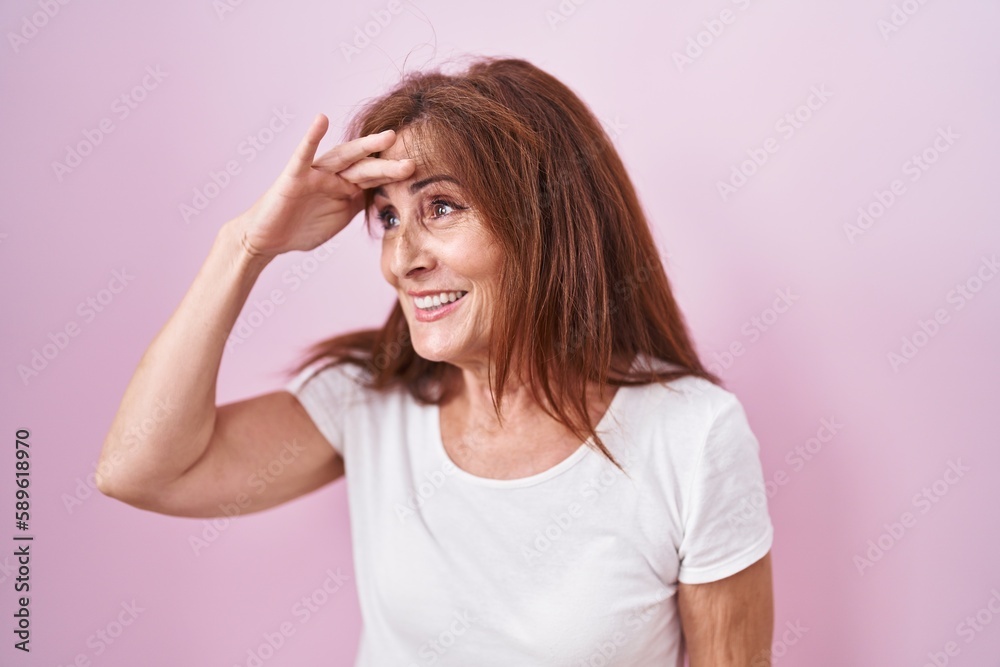 Middle age woman standing over pink background very happy and smiling looking far away with hand over head. searching concept. Middle age woman standing over pink background very happy and smiling looking far away with hand over head. searching concept.