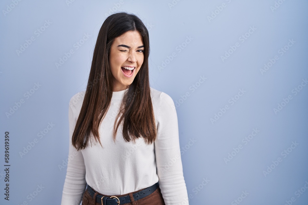 Young brunette woman standing over blue background winking looking at the camera with sexy expression, cheerful and happy face. Young brunette woman standing over blue background winking looking at the camera with sexy expression, cheerful and happy face.