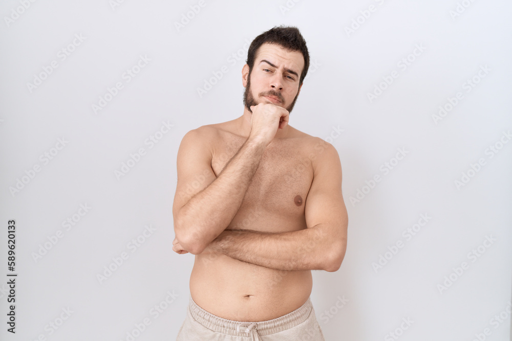 Young hispanic man standing shirtless over white background looking confident at the camera with smile with crossed arms and hand raised on chin. thinking positive.