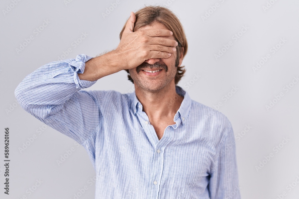 Caucasian man with mustache standing over white background smiling and laughing with hand on face covering eyes for surprise. blind concept. Caucasian man with mustache standing over white background smiling and laughing with hand on face covering eyes for surprise. blind concept.