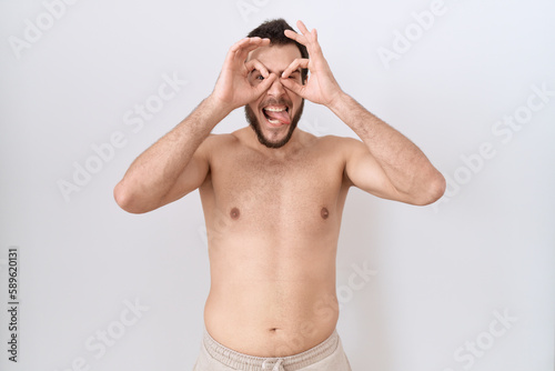 Young hispanic man standing shirtless over white background doing ok gesture like binoculars sticking tongue out, eyes looking through fingers. crazy expression.