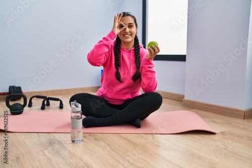 Young hispanic woman sitting on yoga mat eating apple smiling happy doing ok sign with hand on eye looking through fingers