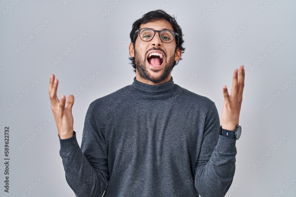 Handsome latin man standing over isolated background celebrating mad and crazy for success with arms raised and closed eyes screaming excited. winner concept Handsome latin man standing over isolated background celebrating mad and crazy for success with arms raised and closed eyes screaming excited. winner concept