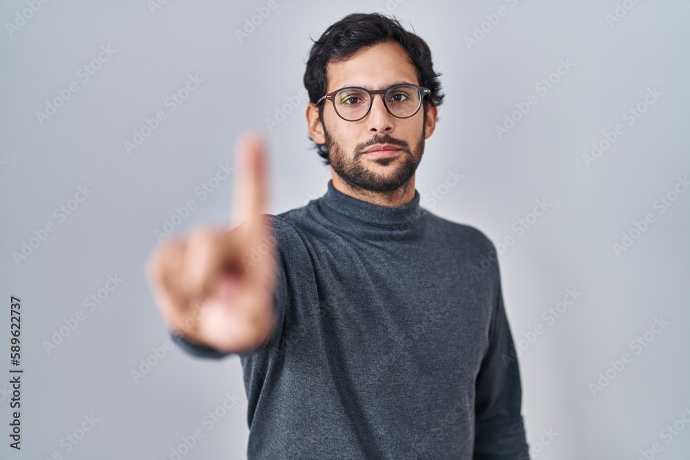 Handsome latin man standing over isolated background pointing with finger up and angry expression, showing no gesture Handsome latin man standing over isolated background pointing with finger up and angry expression, showing no gesture