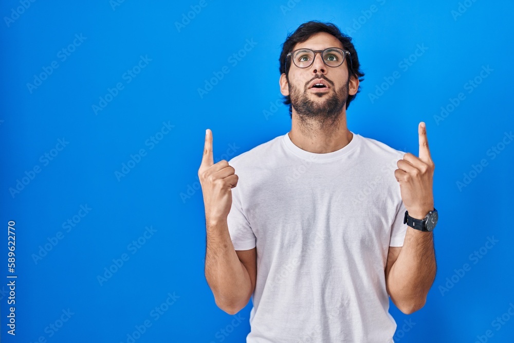Handsome latin man standing over blue background amazed and surprised looking up and pointing with fingers and raised arms. Handsome latin man standing over blue background amazed and surprised looking up and pointing with fingers and raised arms.