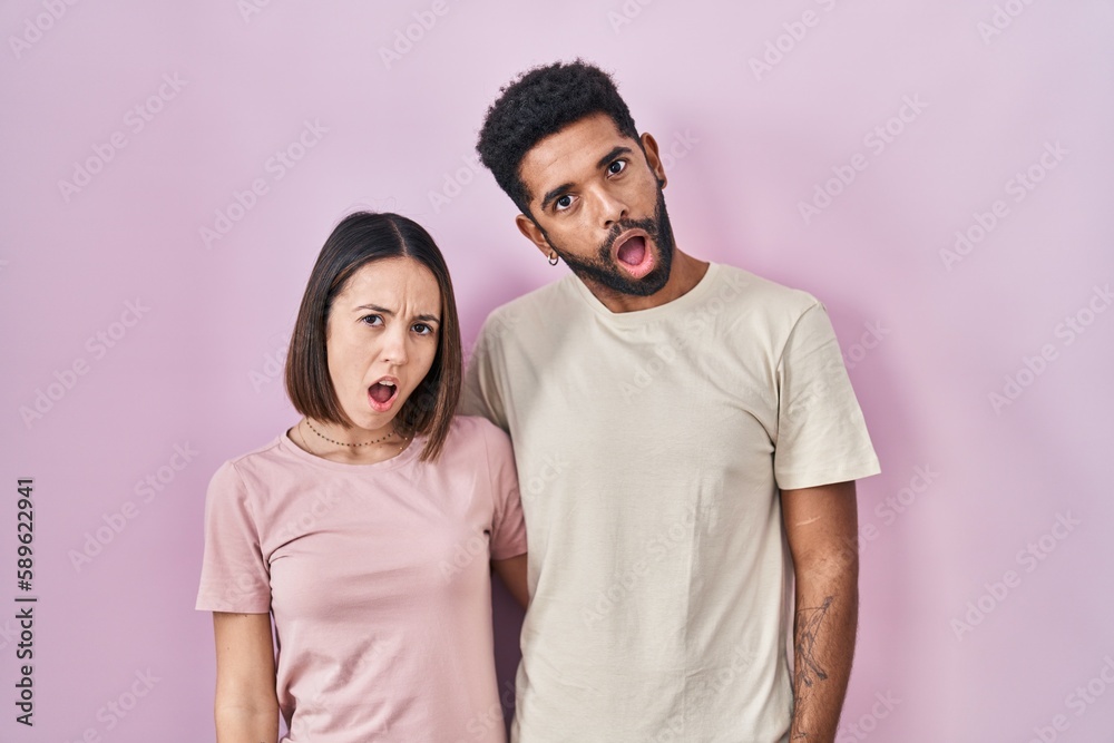 Young hispanic couple together over pink background in shock face, looking skeptical and sarcastic, surprised with open mouth Young hispanic couple together over pink background in shock face, looking skeptical and sarcastic, surprised with open mouth