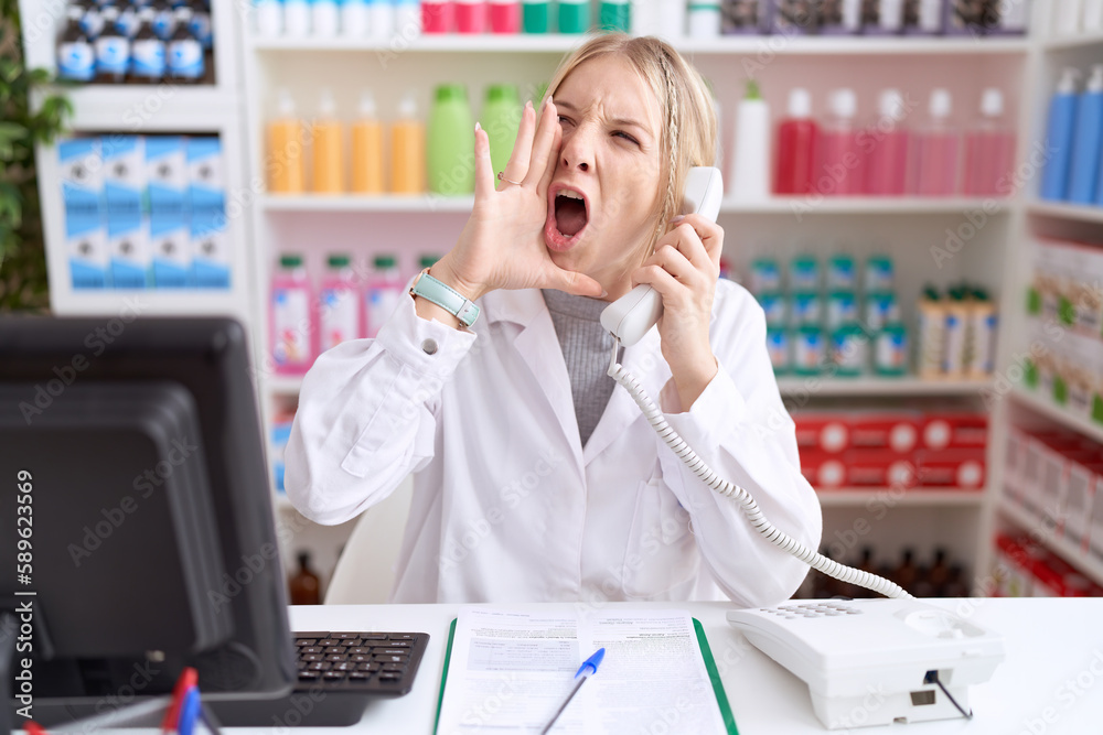 Young caucasian woman working at pharmacy drugstore speaking on the telephone shouting and screaming loud to side with hand on mouth. communication concept.
