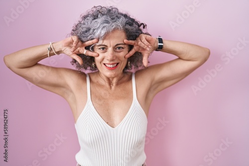 Middle age woman with grey hair standing over pink background doing peace symbol with fingers over face, smiling cheerful showing victory