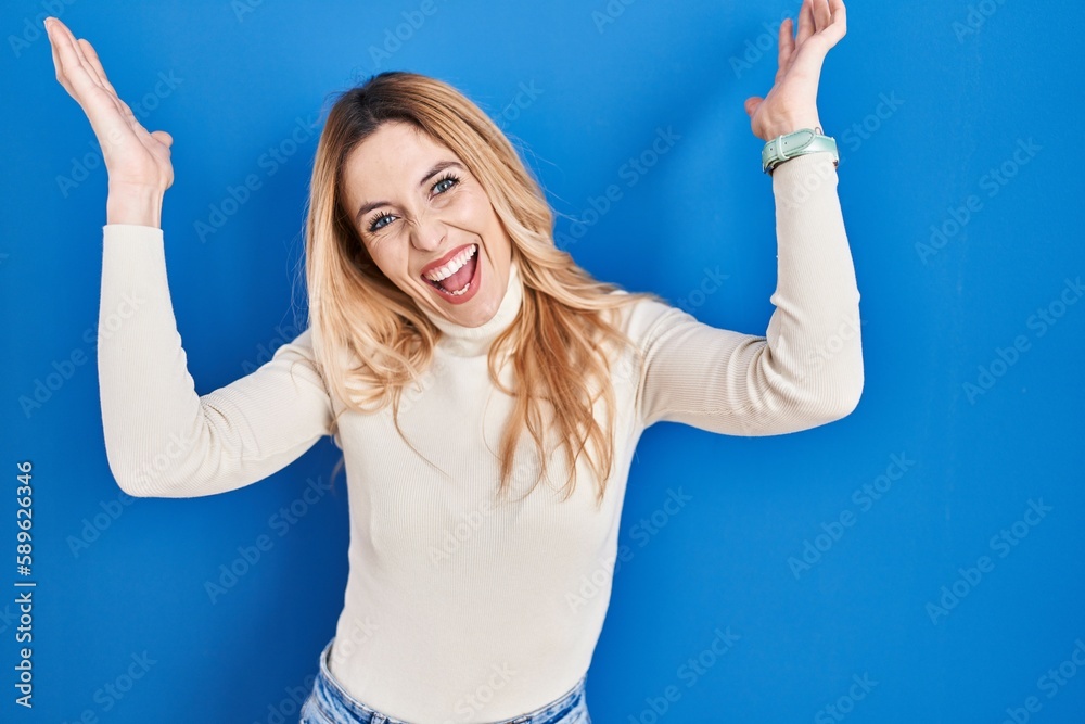 Fototapeta premium Young caucasian woman standing over blue background celebrating crazy and amazed for success with arms raised and open eyes screaming excited. winner concept