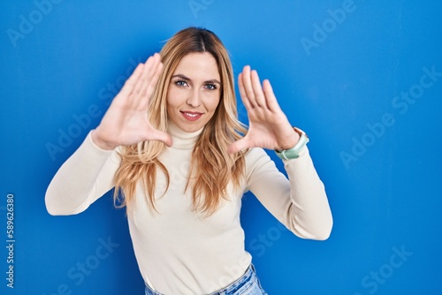 Young caucasian woman standing over blue background doing frame using hands palms and fingers  camera perspective