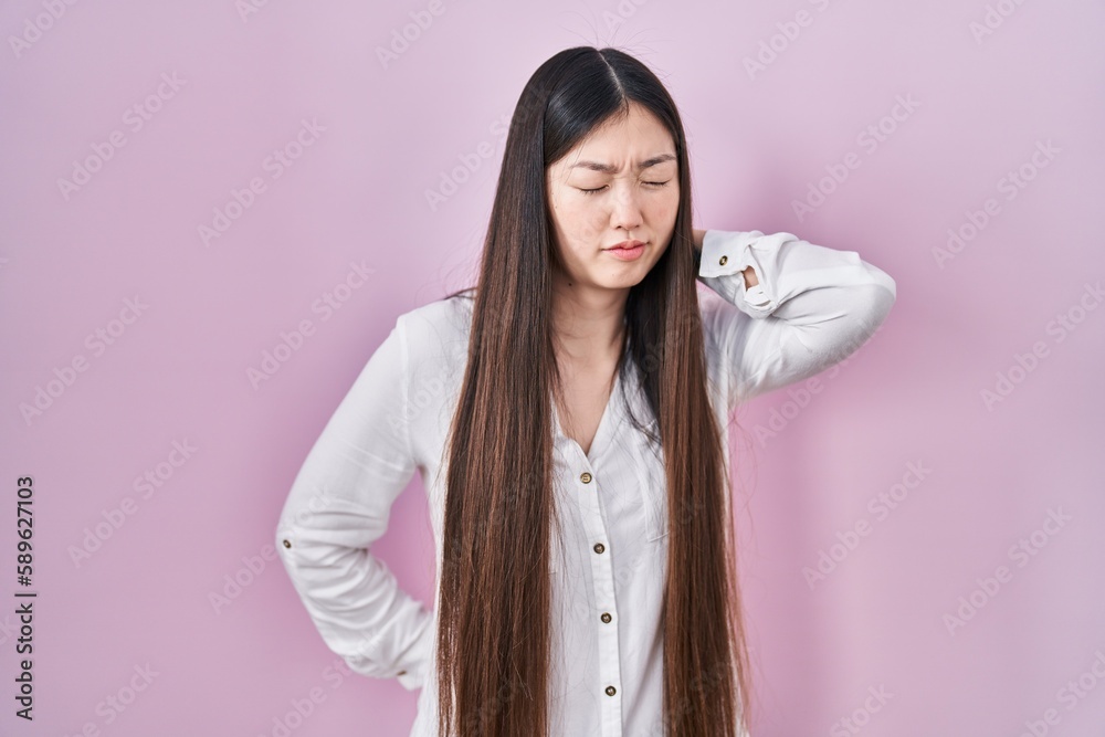 Chinese young woman standing over pink background suffering of neck ache injury, touching neck with hand, muscular pain