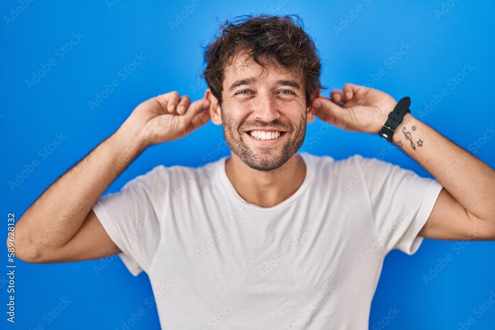 Hispanic young man standing over blue background smiling pulling ears with fingers, funny gesture. audition problem Hispanic young man standing over blue background smiling pulling ears with fingers, funny gesture. audition problem
