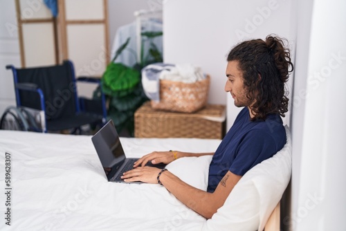 Young hispanic man disabled using laptop sitting on bed at bedroom