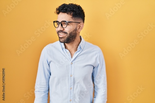 Hispanic man with beard standing over yellow background looking away to side with smile on face, natural expression. laughing confident.