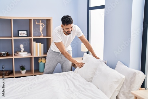 Young hispanic man make bed standing at bedroom