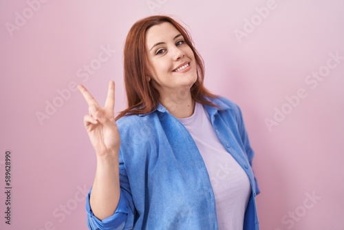 Young hispanic woman with red hair standing over pink background smiling looking to the camera showing fingers doing victory sign. number two. © Krakenimages.com