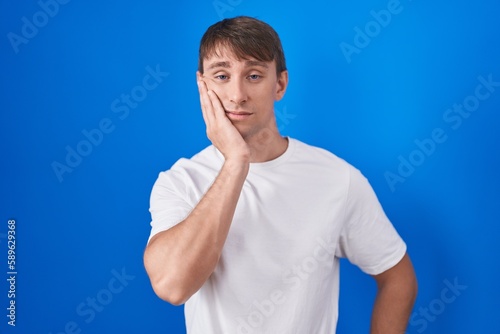 Caucasian blond man standing over blue background thinking looking tired and bored with depression problems with crossed arms.