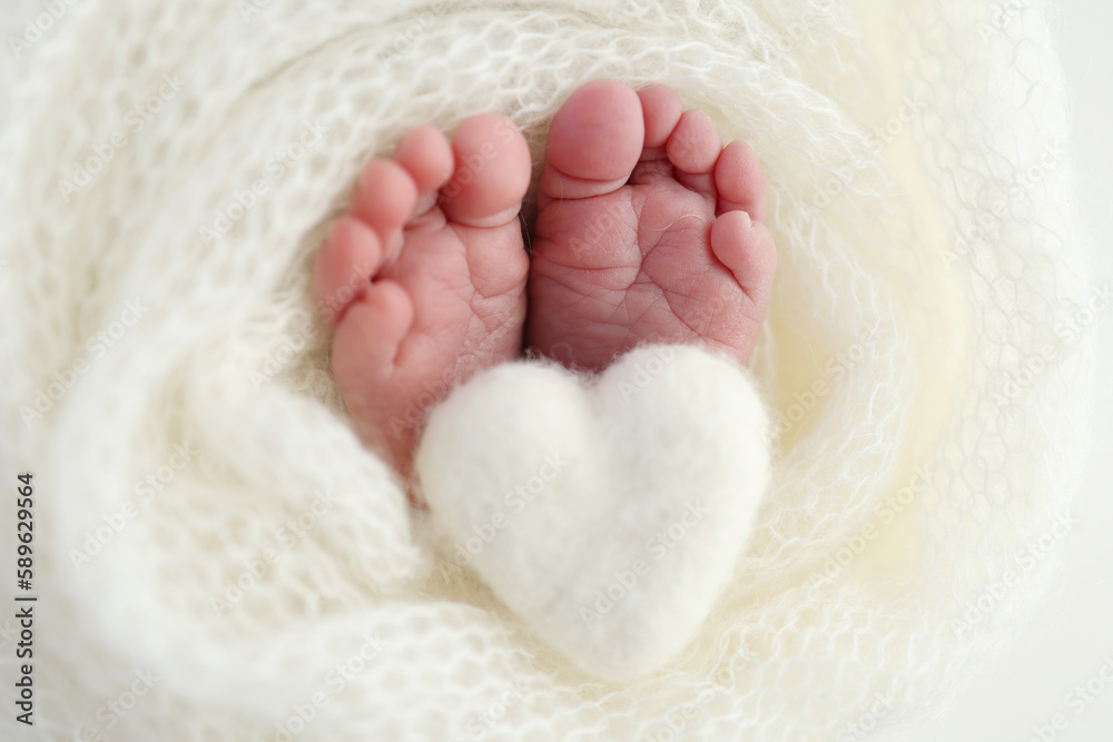 Knitted white heart in the legs of a baby. Macro photography. The tiny foot of a newborn baby. Soft feet of a new born in a white wool blanket. Close up of toes, heels and feet of a newborn. Knitted white heart in the legs of a baby. Macro photography. The tiny foot of a newborn baby. Soft feet of a new born in a white wool blanket. Close up of toes, heels and feet of a newborn.