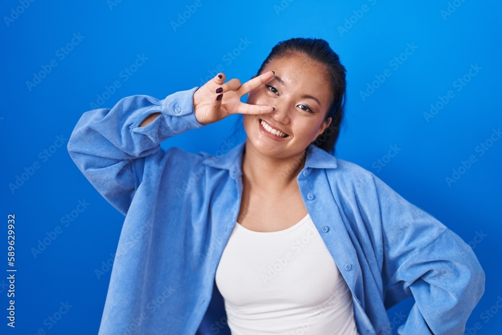 Asian young woman standing over blue background doing peace symbol with fingers over face, smiling cheerful showing victory Asian young woman standing over blue background doing peace symbol with fingers over face, smiling cheerful showing victory