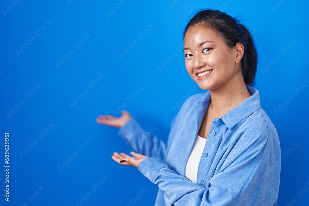 Asian young woman standing over blue background inviting to enter smiling natural with open hand Asian young woman standing over blue background inviting to enter smiling natural with open hand