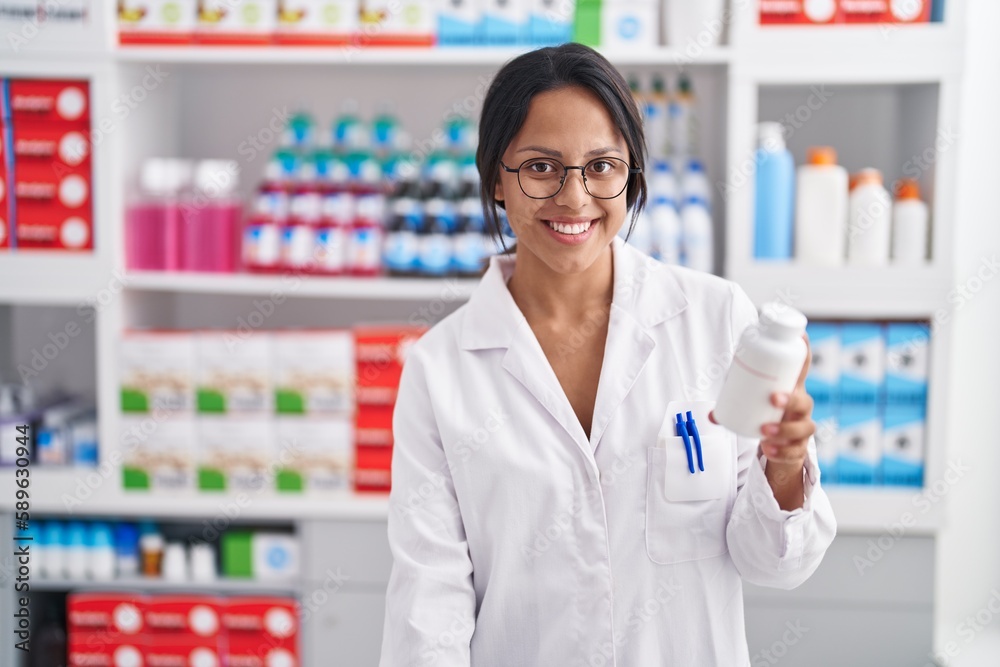 Young hispanic woman pharmacist smiling confident holding pills bottle at pharmacy