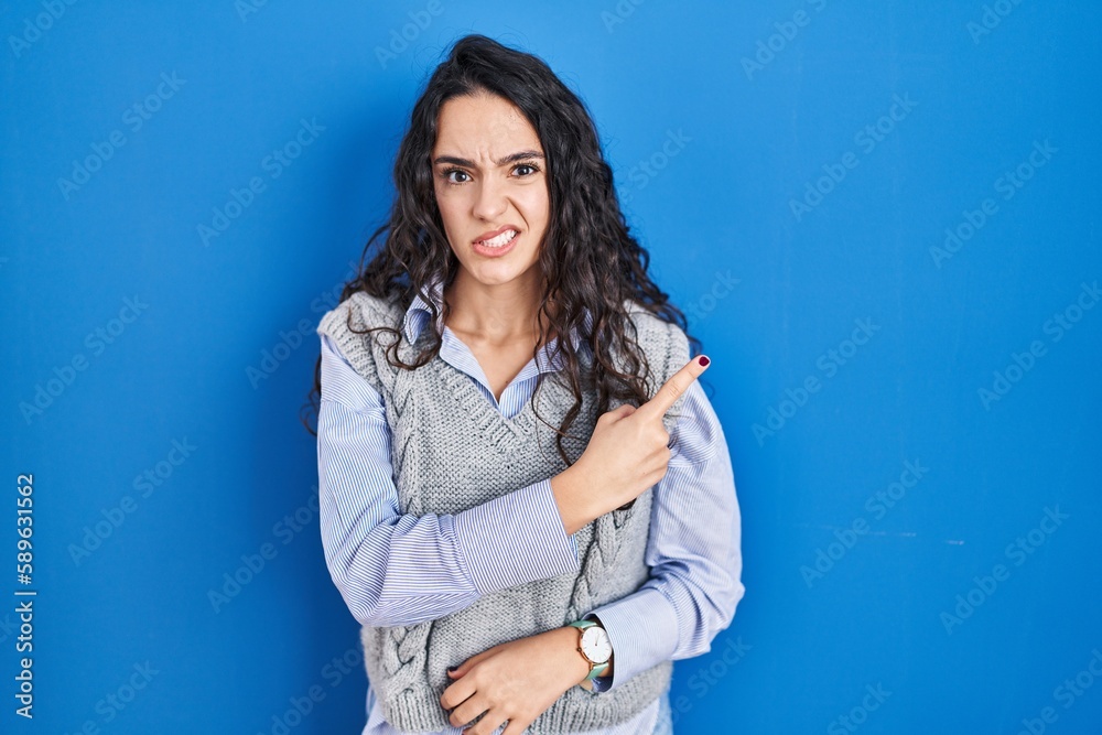 Young brunette woman standing over blue background pointing aside worried and nervous with forefinger, concerned and surprised expression Young brunette woman standing over blue background pointing aside worried and nervous with forefinger, concerned and surprised expression