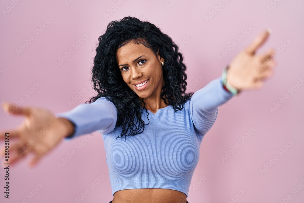 Middle age hispanic woman standing over pink background looking at the camera smiling with open arms for hug. cheerful expression embracing happiness.