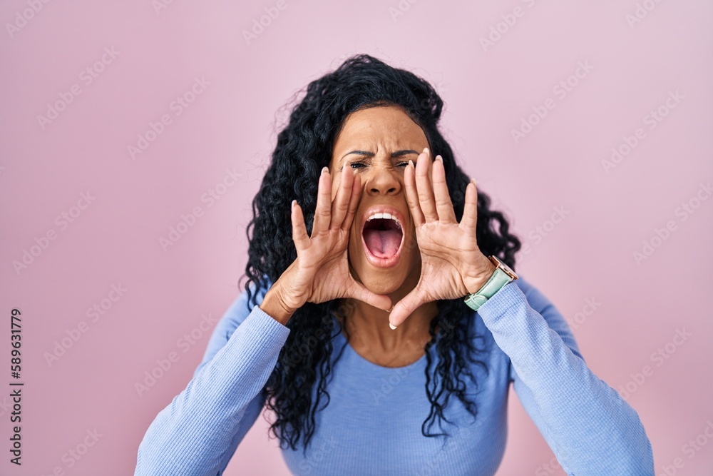 Middle age hispanic woman standing over pink background shouting angry out loud with hands over mouth Middle age hispanic woman standing over pink background shouting angry out loud with hands over mouth