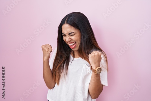 Young arab woman standing over pink background very happy and excited doing winner gesture with arms raised  smiling and screaming for success. celebration concept.