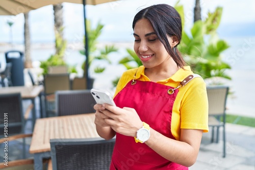 Young beautiful arab woman waitress smiling confident using smartphone at coffee shop terrace
