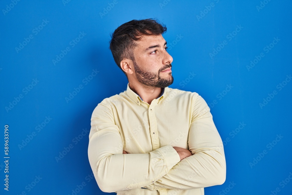 Handsome hispanic man standing over blue background looking to the side with arms crossed convinced and confident