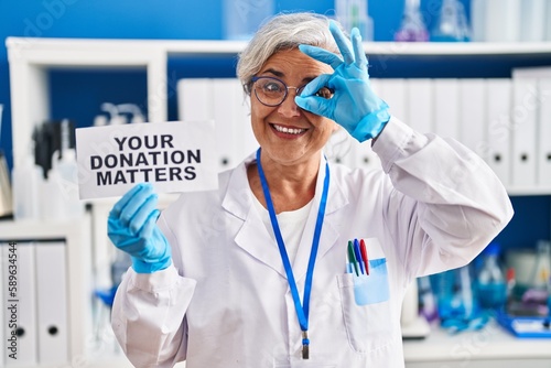 Middle age woman with grey hair working at scientist laboratory holding your donation matters banner smiling happy doing ok sign with hand on eye looking through fingers