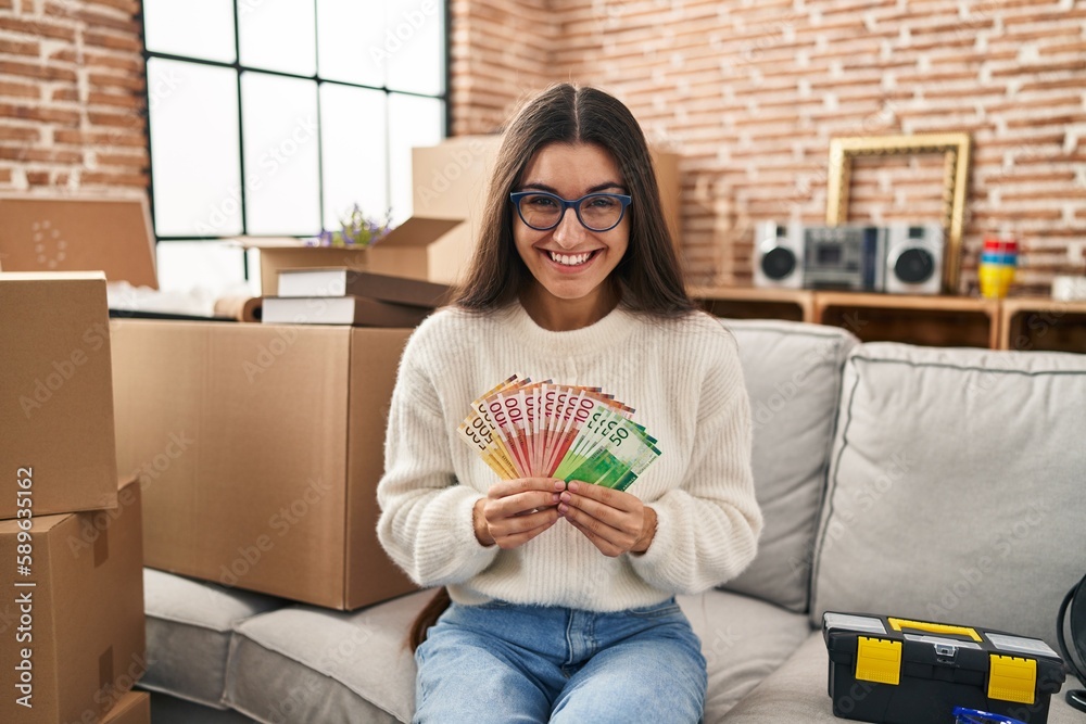 Young hispanic woman sitting on the sofa at new home holding money smiling and laughing hard out loud because funny crazy joke. Young hispanic woman sitting on the sofa at new home holding money smiling and laughing hard out loud because funny crazy joke.