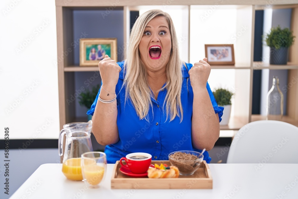 Caucasian plus size woman eating breakfast at home celebrating surprised and amazed for success with arms raised and open eyes. winner concept.