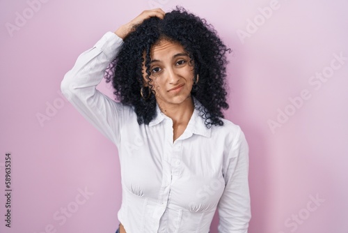 Hispanic woman with curly hair standing over pink background confuse and wondering about question. uncertain with doubt, thinking with hand on head. pensive concept.