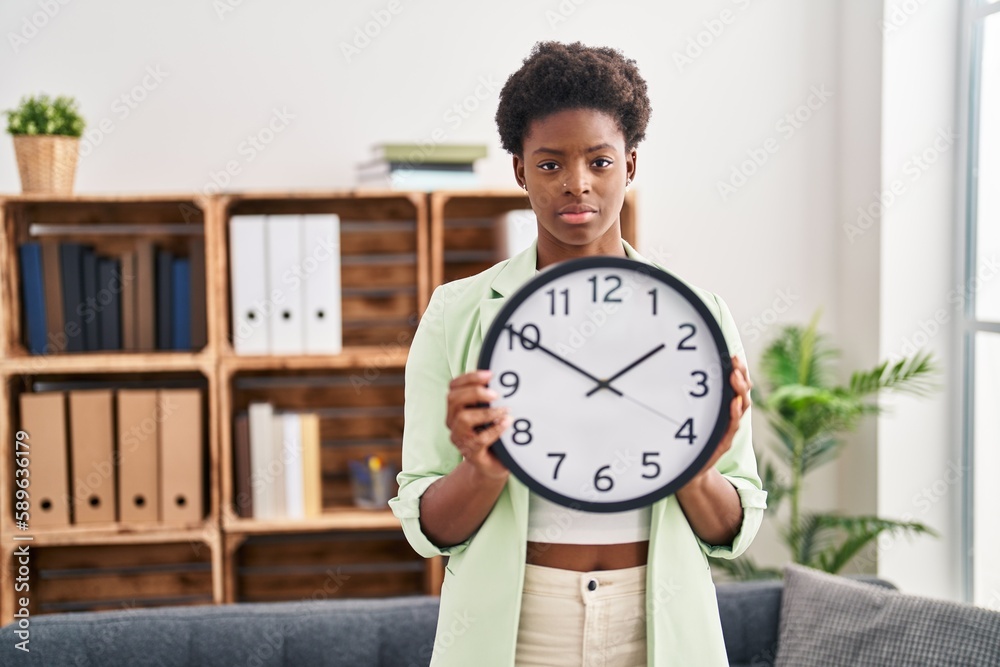 African american woman holding big clock relaxed with serious expression on face. simple and natural looking at the camera. African american woman holding big clock relaxed with serious expression on face. simple and natural looking at the camera.